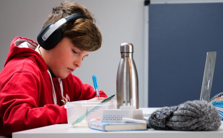 A boy wearing headphones writing at a desk. On the desk is a laptop and a water bottle and hat.