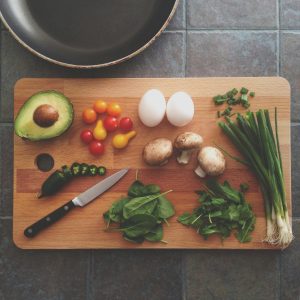 Wooden board with a knife, tomatoes, eggs, avocado spring onion and herbs. A frying pan is above the board on the left
